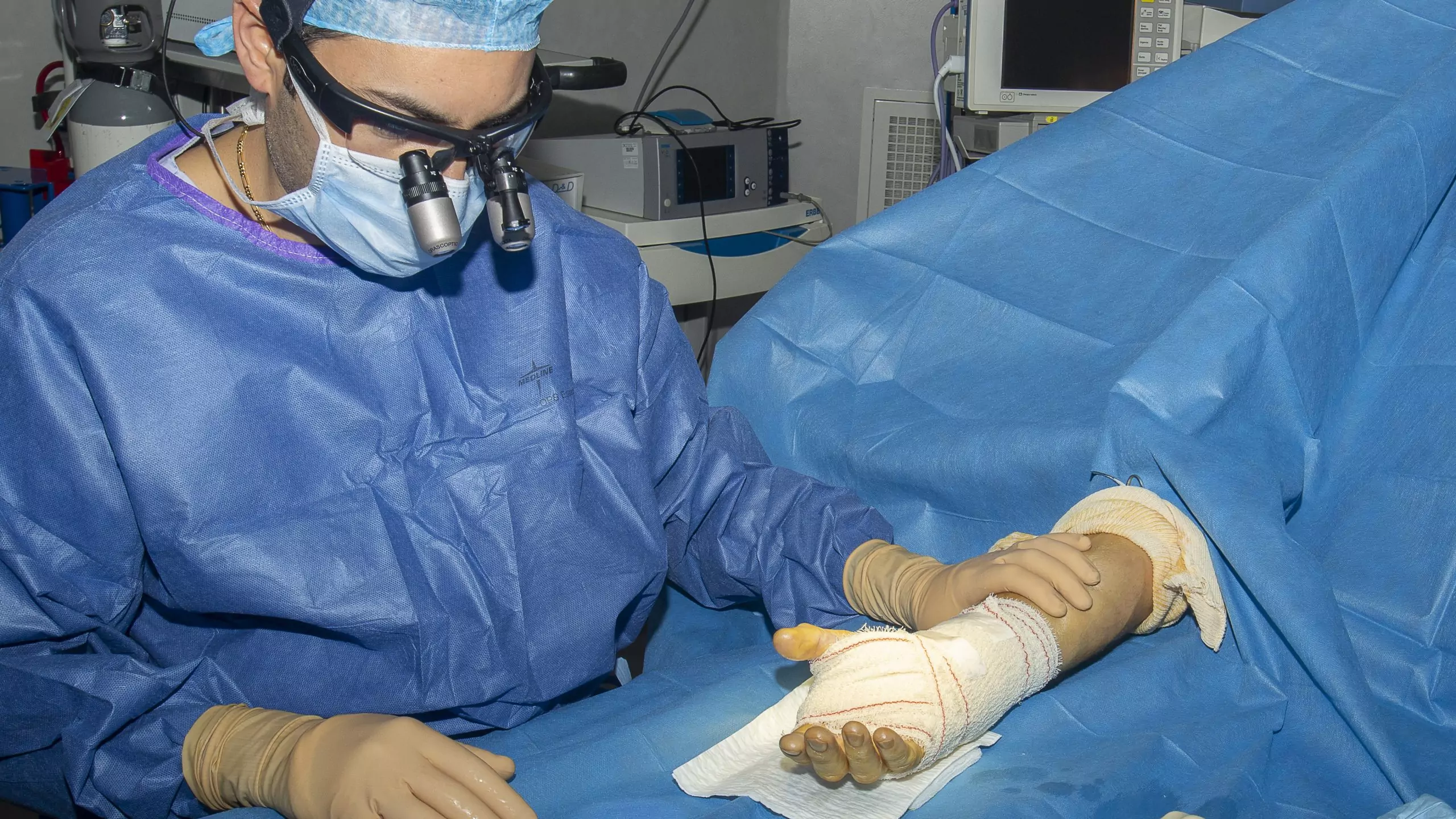 Chirurgien opérant une main bandée, équipé de lunettes loupes et de gants stériles en bloc opératoire.